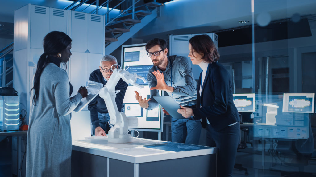 Engineers Meeting in Robotic Research Laboratory: Engineers, Scientists and Developers Gathered Around Illuminated Conference Table, Talking, Using Tablet and Analysing Design of Industrial Robot Arm